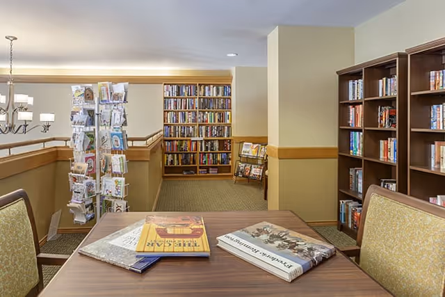 Library-style common area with a wooden table and chairs in the foreground, bookshelves lining the walls, and a rotating greeting card rack.