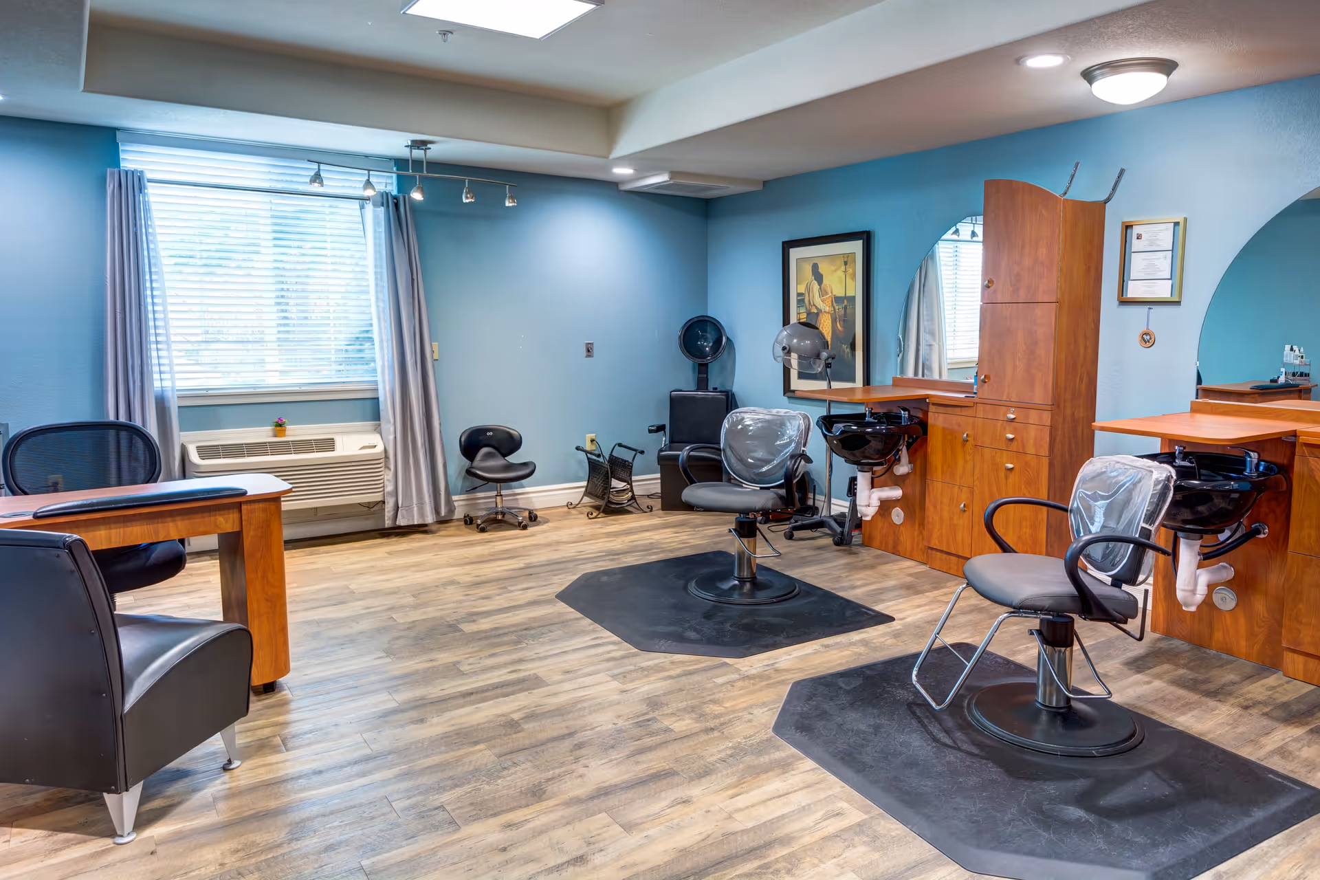 Interior view of a senior living facility's hair salon with two salon chairs in front of mirrors and wash basins, a desk with chairs, light blue walls, wooden flooring, and a window with curtains.