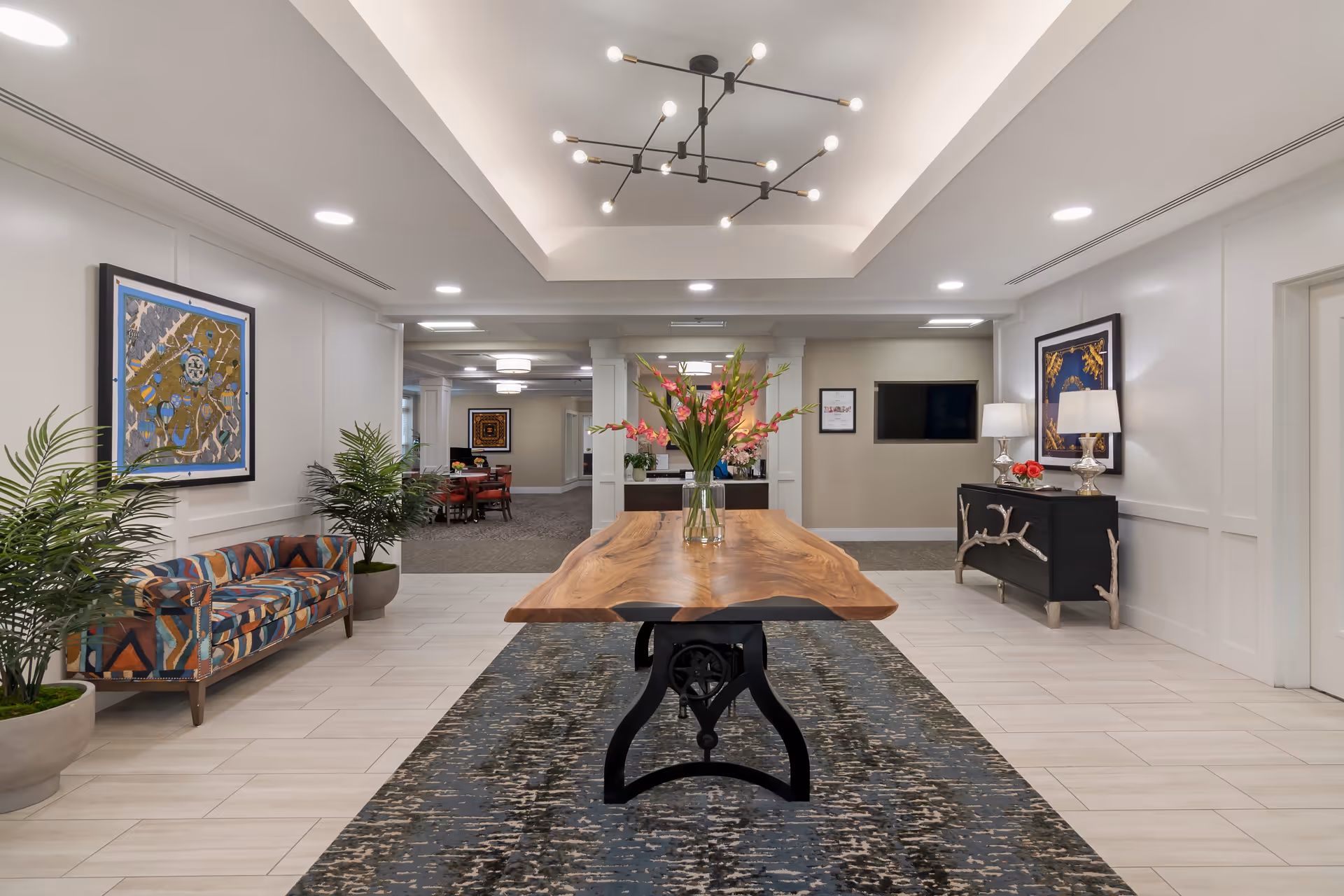 A spacious and well-lit interior common area featuring a large wooden table with a glass vase of pink flowers in the center. The room has a modern chandelier hanging from a tray ceiling, patterned couch on the left, two potted plants, framed artwork on the walls, and a black sideboard with two lamps and decorative items on the right. In the background, there is a dining area with tables and chairs.