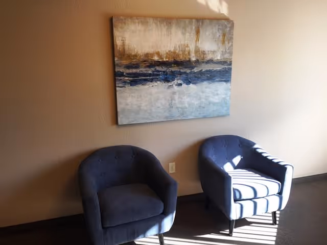 Two blue upholstered armchairs sit beneath an abstract painting on a sunlit beige wall.