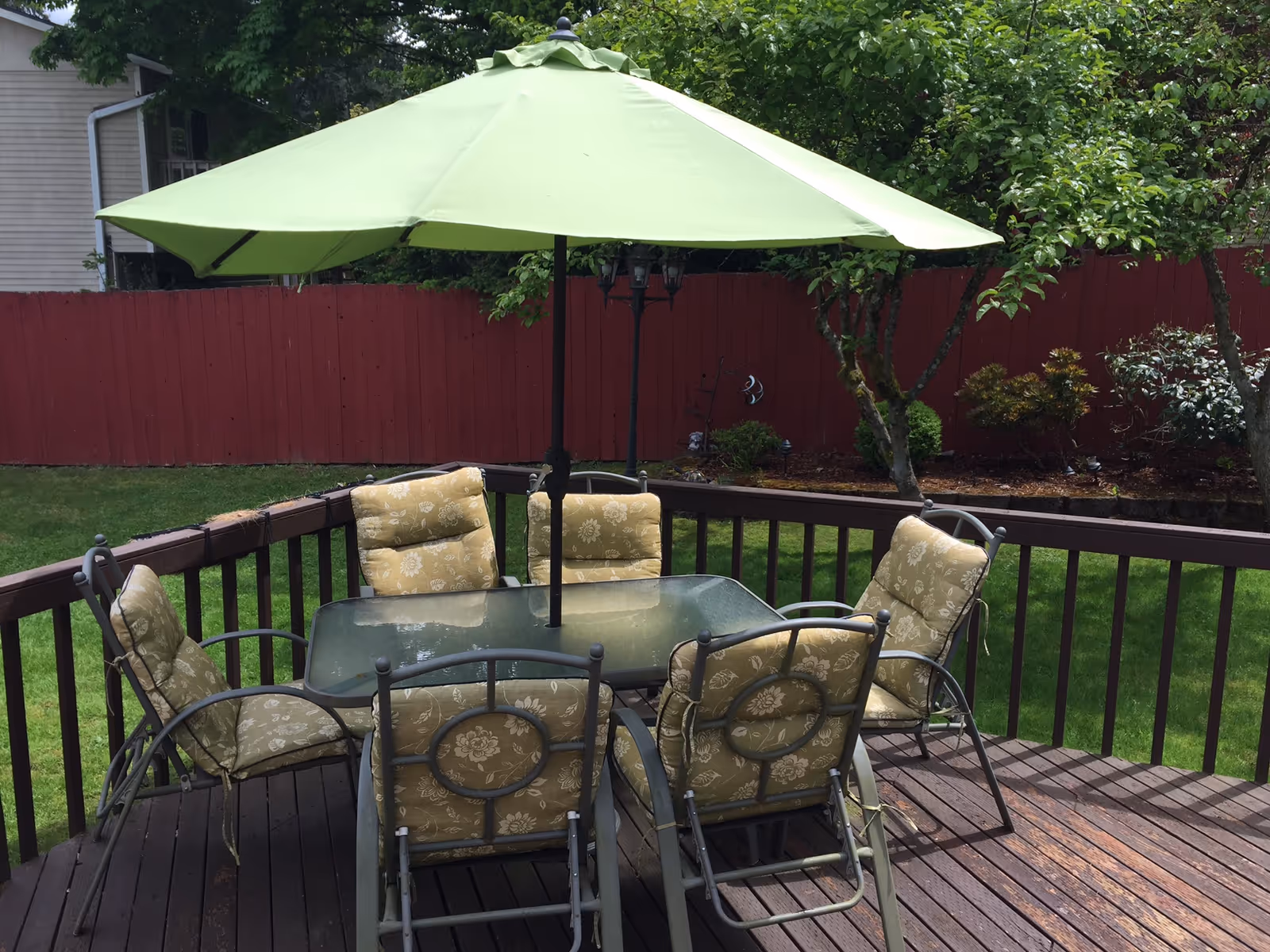 Outdoor wooden deck with a glass-top table surrounded by six cushioned chairs with floral patterns. A large green patio umbrella is centered over the table. The deck overlooks a grassy yard with a red wooden fence and some trees and shrubs in the background.
