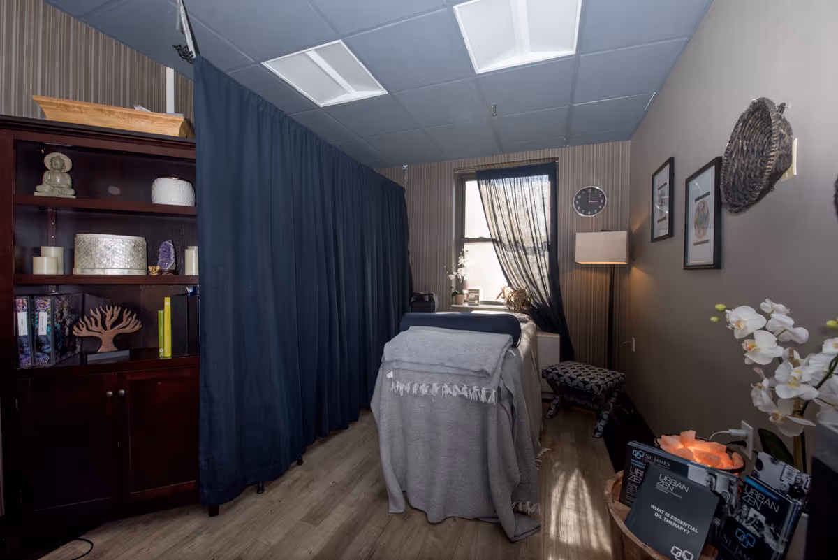 A cozy therapy or massage room with a treatment table covered in gray blankets, a dark curtain divider, a wooden cabinet with decorative items, a window with sheer black curtains, a floor lamp, wall art, and a small stool. There are also informational brochures and a salt lamp on a small table.