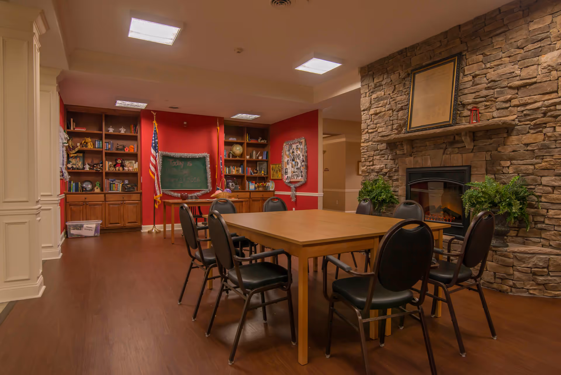 A cozy common room with a wooden table surrounded by eight black chairs. The room features a stone fireplace with a framed picture above it and two potted plants on either side. In the background, there are built-in wooden shelves filled with books and decorative items, an American flag, and a chalkboard with writing on it. The walls are painted red and beige, and the floor is wooden.
