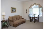 A cozy living room with a brown patterned sofa, a wooden side table with a lamp and a plant, a small round dining table with two chairs near a window with decorative curtains, and a framed picture on the wall.