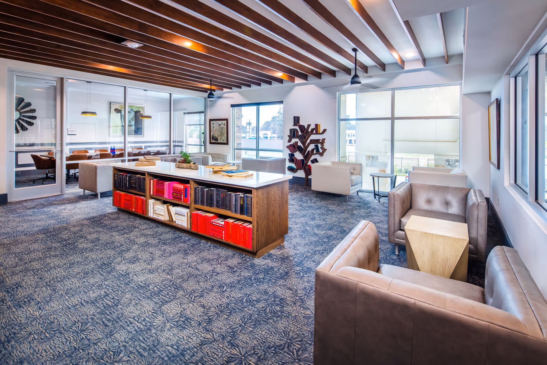A modern and spacious senior community lounge area with comfortable leather armchairs arranged around small wooden tables. In the center, there is a large wooden bookshelf with a white countertop, filled with colorful books. The room features large windows allowing natural light to fill the space, a patterned carpet, and a ceiling with wooden beams and recessed lighting. A glass-walled conference room with a long table and chairs is visible in the background.