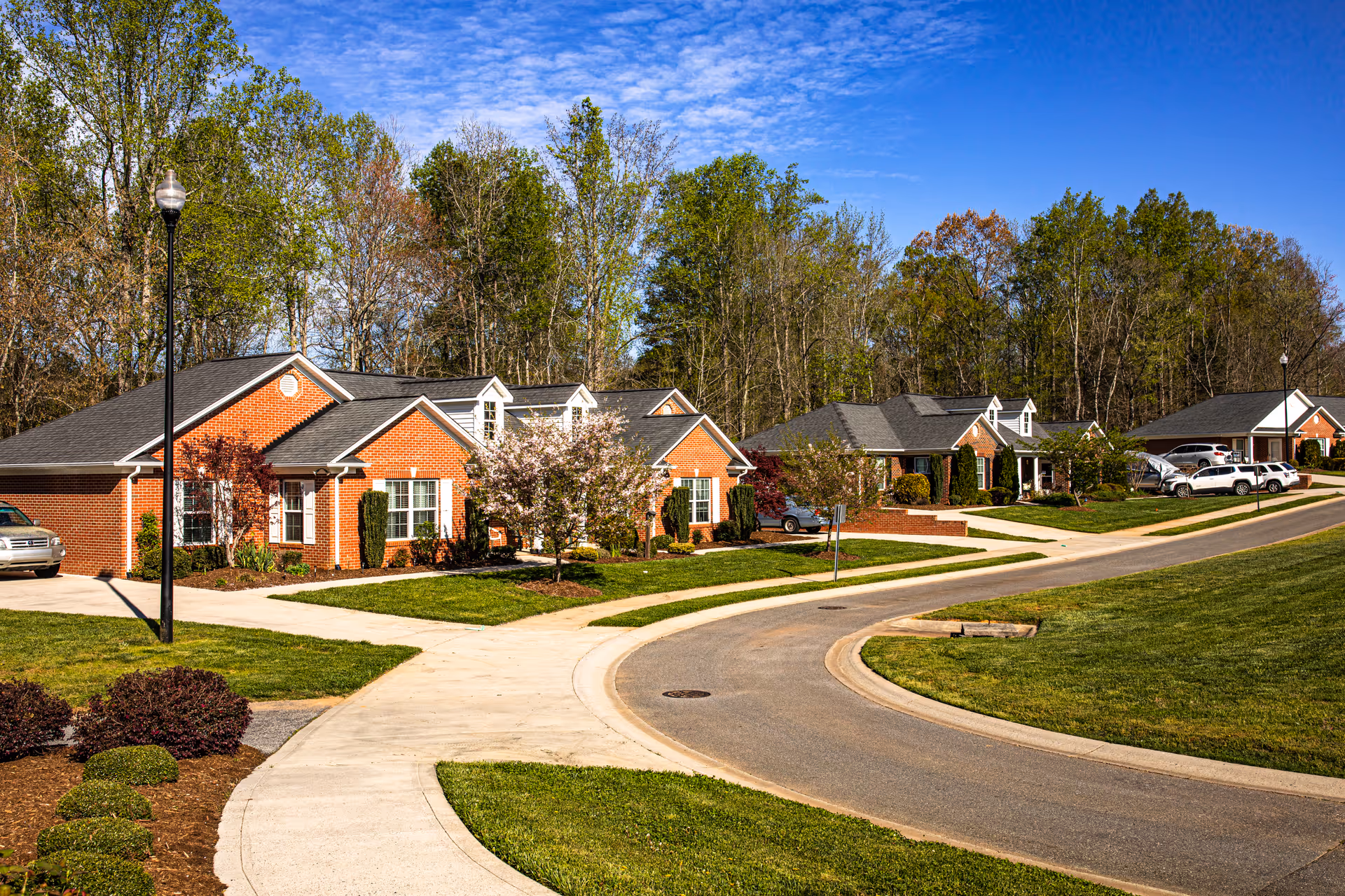 A curved street lined with single-story brick cottages with well-maintained lawns and small trees, set against a backdrop of tall trees under a blue sky with scattered clouds.