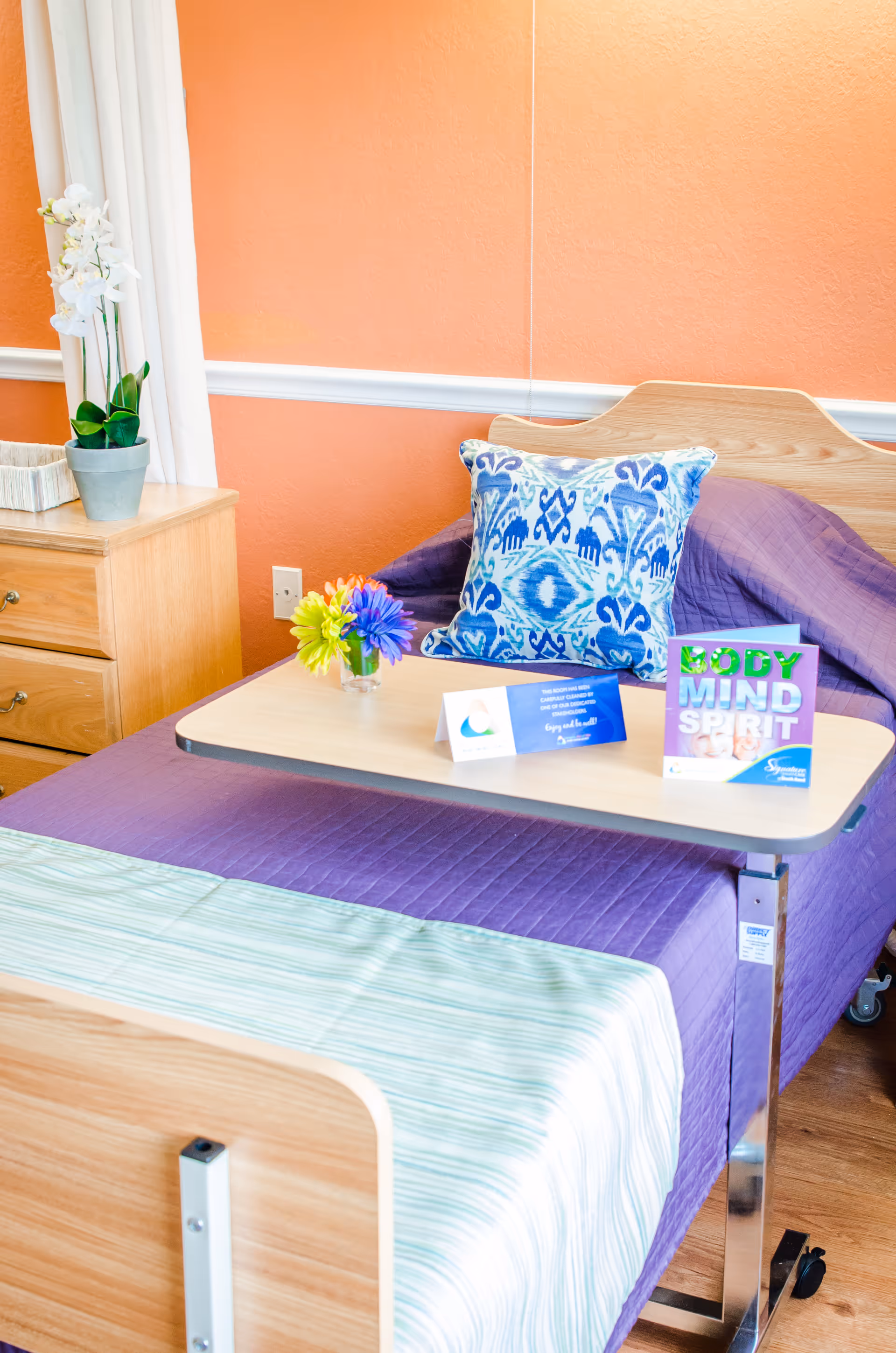 A neatly made bed with a purple quilt and a blue patterned pillow in a room with orange walls. A wooden bedside table with a potted white orchid and a small basket is next to the bed. A wooden overbed table holds a small vase with colorful flowers and two informational cards, one titled 'BODY MIND SPIRIT'.