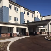 Exterior view of a multi-story senior living facility building with white and blue siding, brick accents, and a covered entrance driveway under a clear blue sky.
