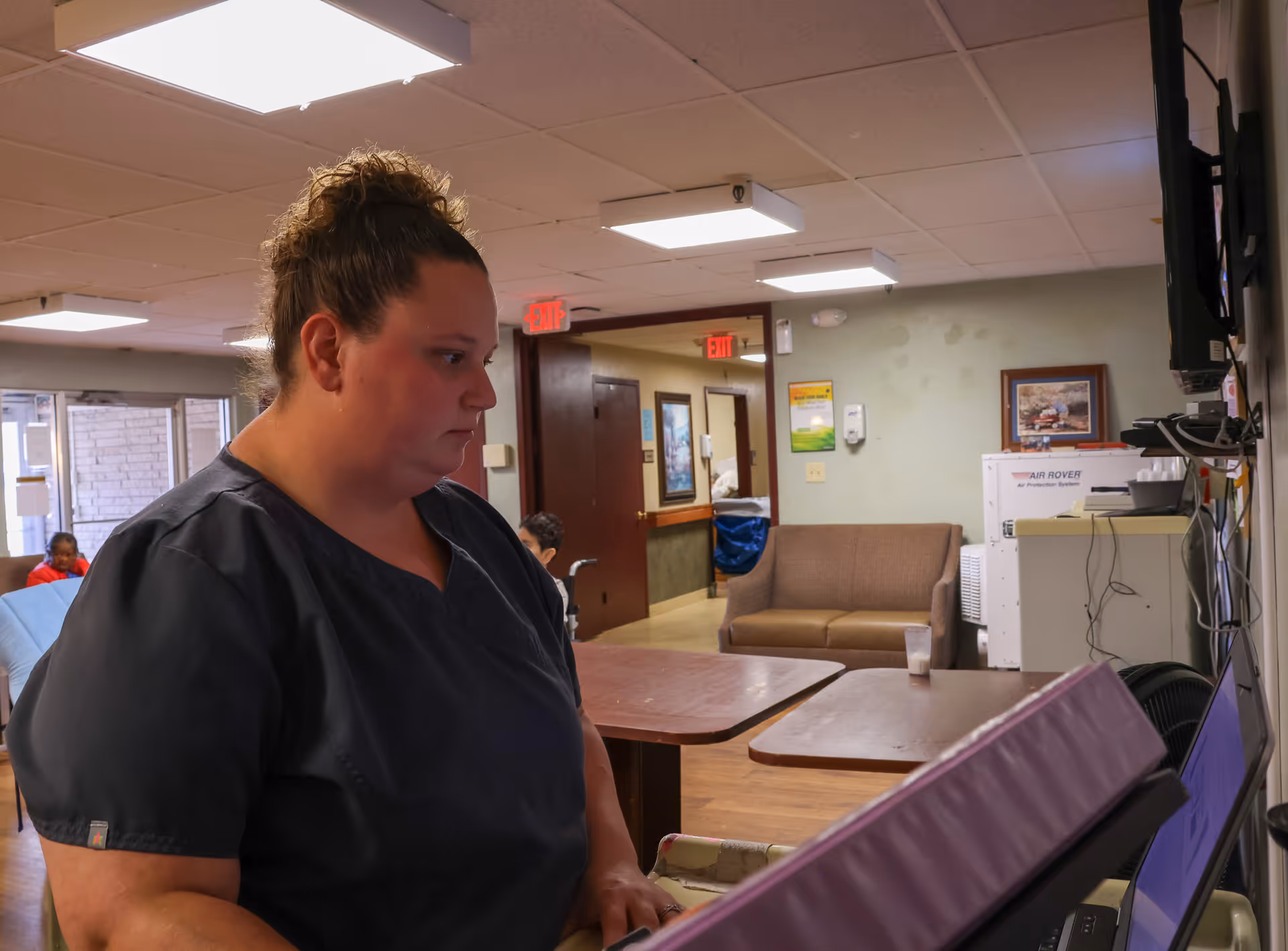 A woman in dark scrubs is working on a computer in a room with tables, a brown couch, and medical equipment. The room has overhead fluorescent lighting and exit signs above doorways in the background.