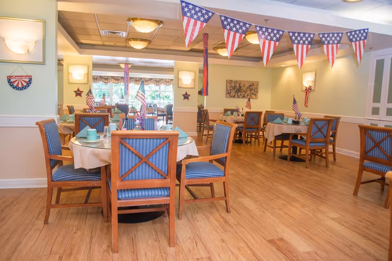 A dining room decorated with American flag-themed bunting and small flags on tables. The room features wooden floors, round tables covered with beige tablecloths, and wooden chairs with blue striped cushions. Wall sconces provide lighting, and there is a painting on one wall. The room has a bright and welcoming atmosphere.