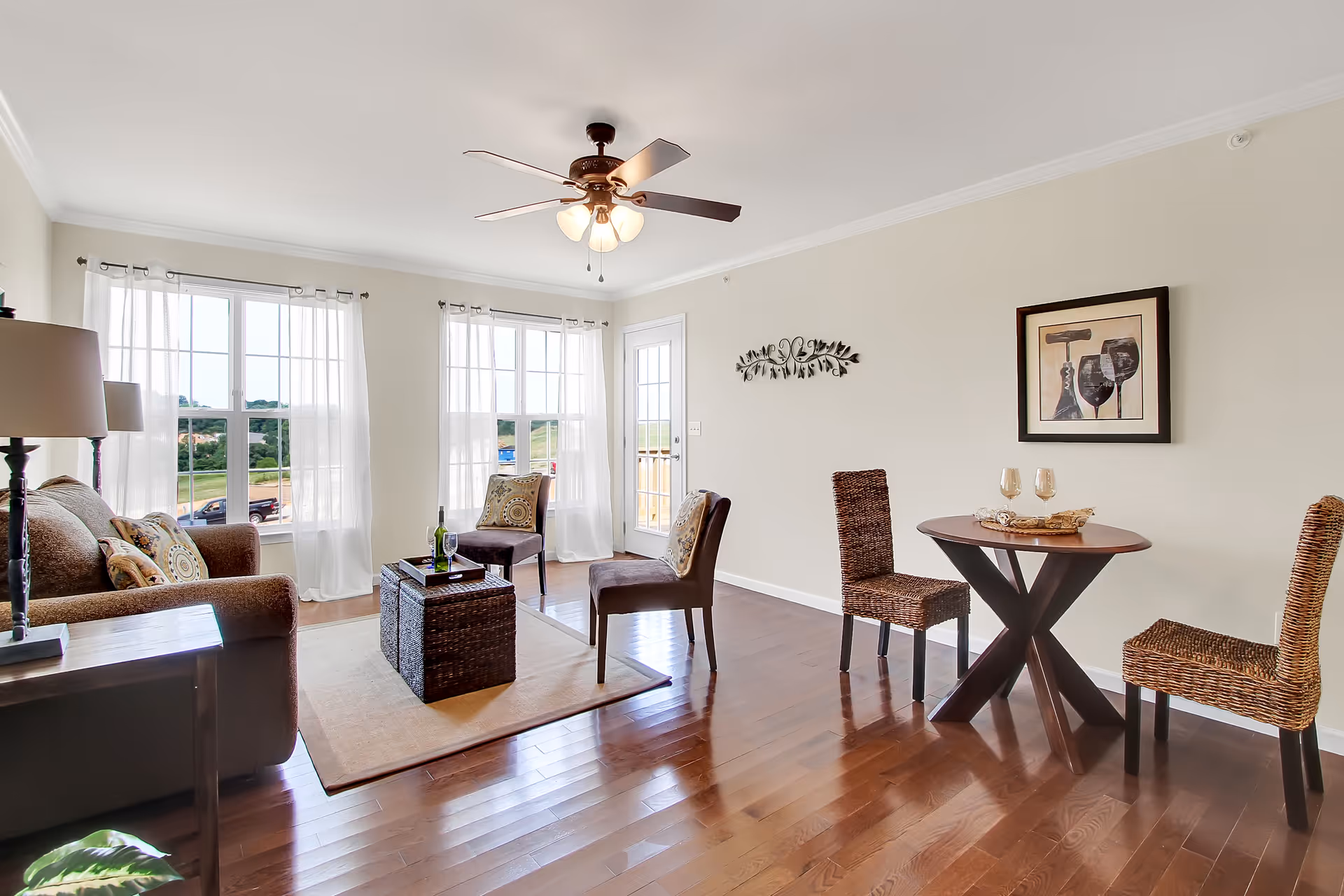A bright living room with large windows covered by sheer white curtains, a ceiling fan with lights, a brown sofa with patterned cushions, two brown chairs, a woven coffee table on a beige rug, and a small round dining table with two woven chairs. A framed picture and a decorative wall piece hang on the light-colored walls.