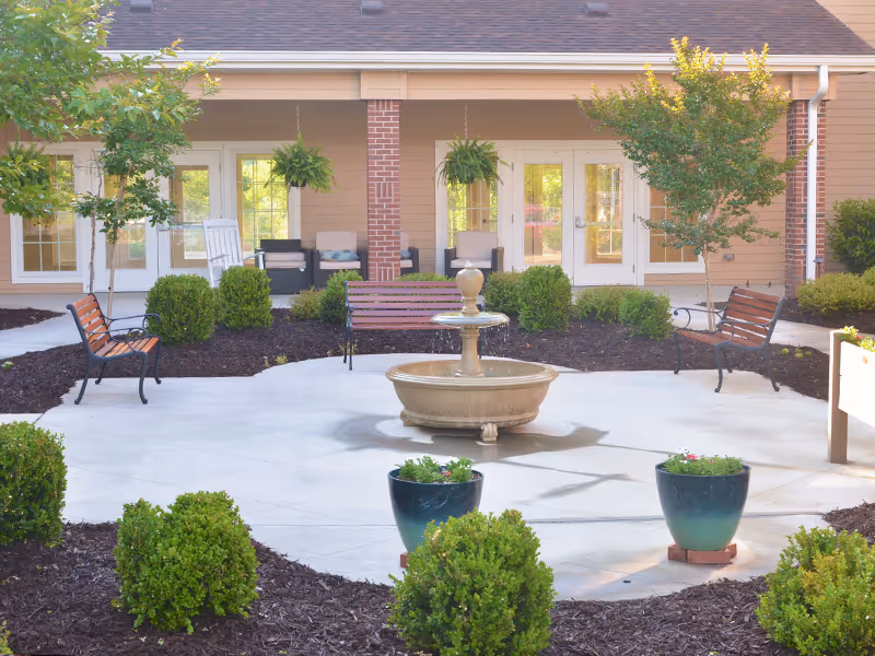 Outdoor courtyard area with a central water fountain surrounded by benches, potted plants, small trees, and bushes. The background shows a building with multiple glass doors and a covered porch with seating.