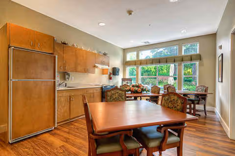 A bright and spacious room with wooden flooring featuring a kitchen area with wooden cabinets and a refrigerator on the left side. In the foreground, there is a wooden dining table with four chairs. In the background, there is a seating area with armchairs and a large window letting in natural light and showing greenery outside.