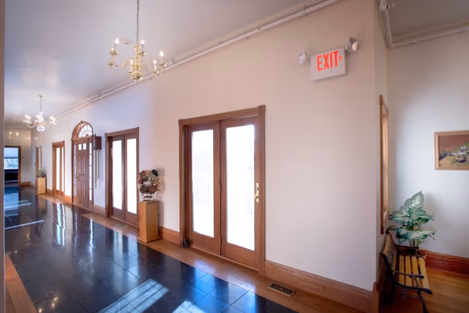 Bright hallway in a senior living facility with large wooden doors and windows letting in natural light. The floor is tiled in black with wooden trim, and there are chandeliers hanging from the ceiling. A wooden bench with a potted plant and a framed picture is visible on the right side. An exit sign is mounted on the wall above the doors.