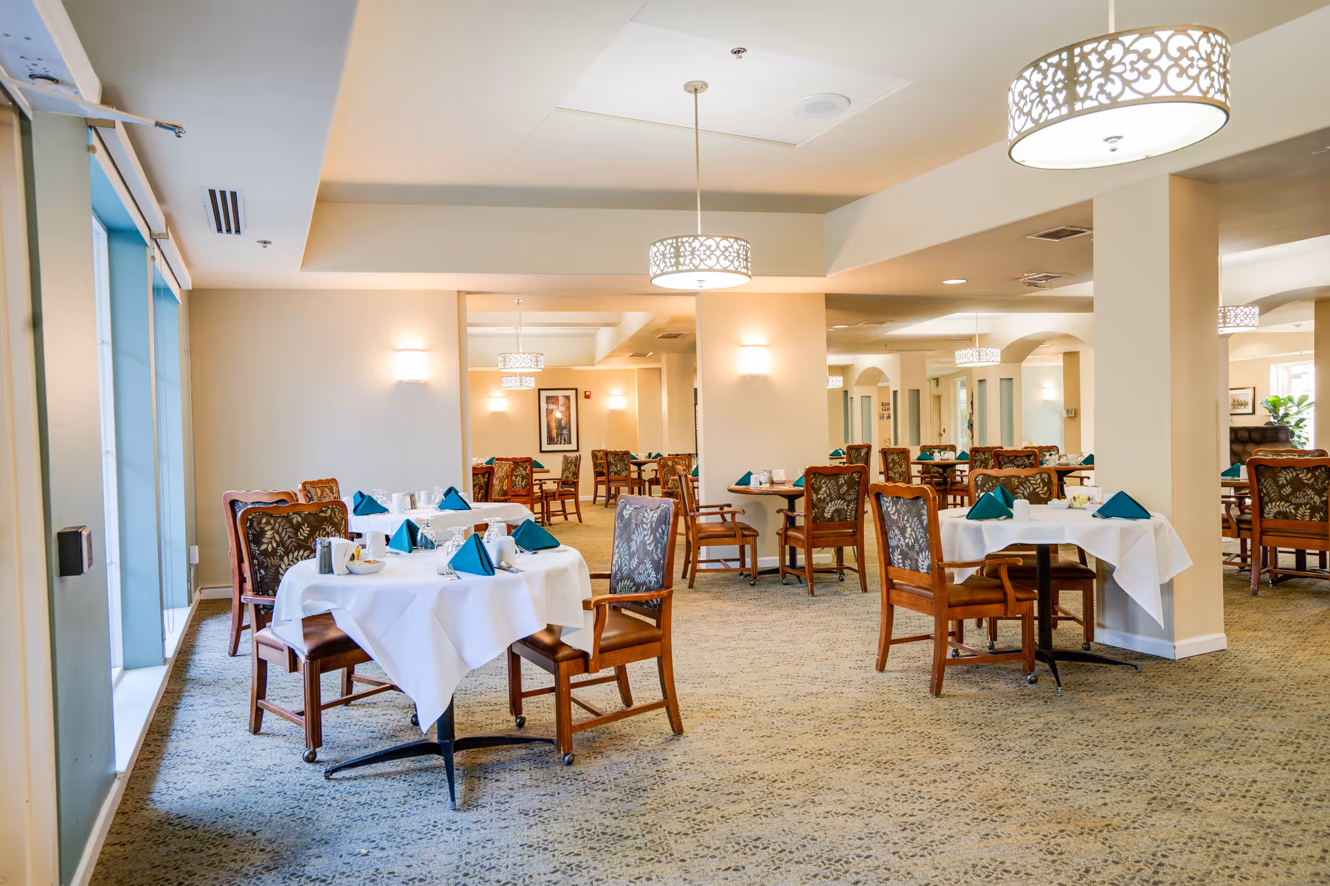 A bright and spacious dining room with multiple round tables covered in white tablecloths, each set with teal folded napkins, cups, and utensils. The room features patterned carpet, wooden chairs with floral upholstery, large windows on the left side, and decorative hanging light fixtures.