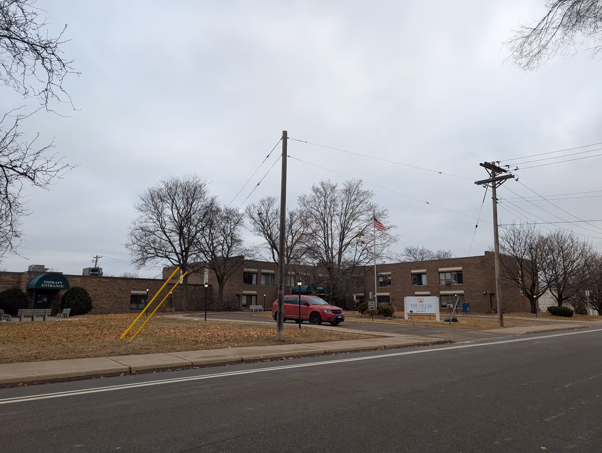 Front exterior of a low two-story brick senior living building with a flagpole, leafless trees, a sign reading "The Villas", and a red SUV parked in front.