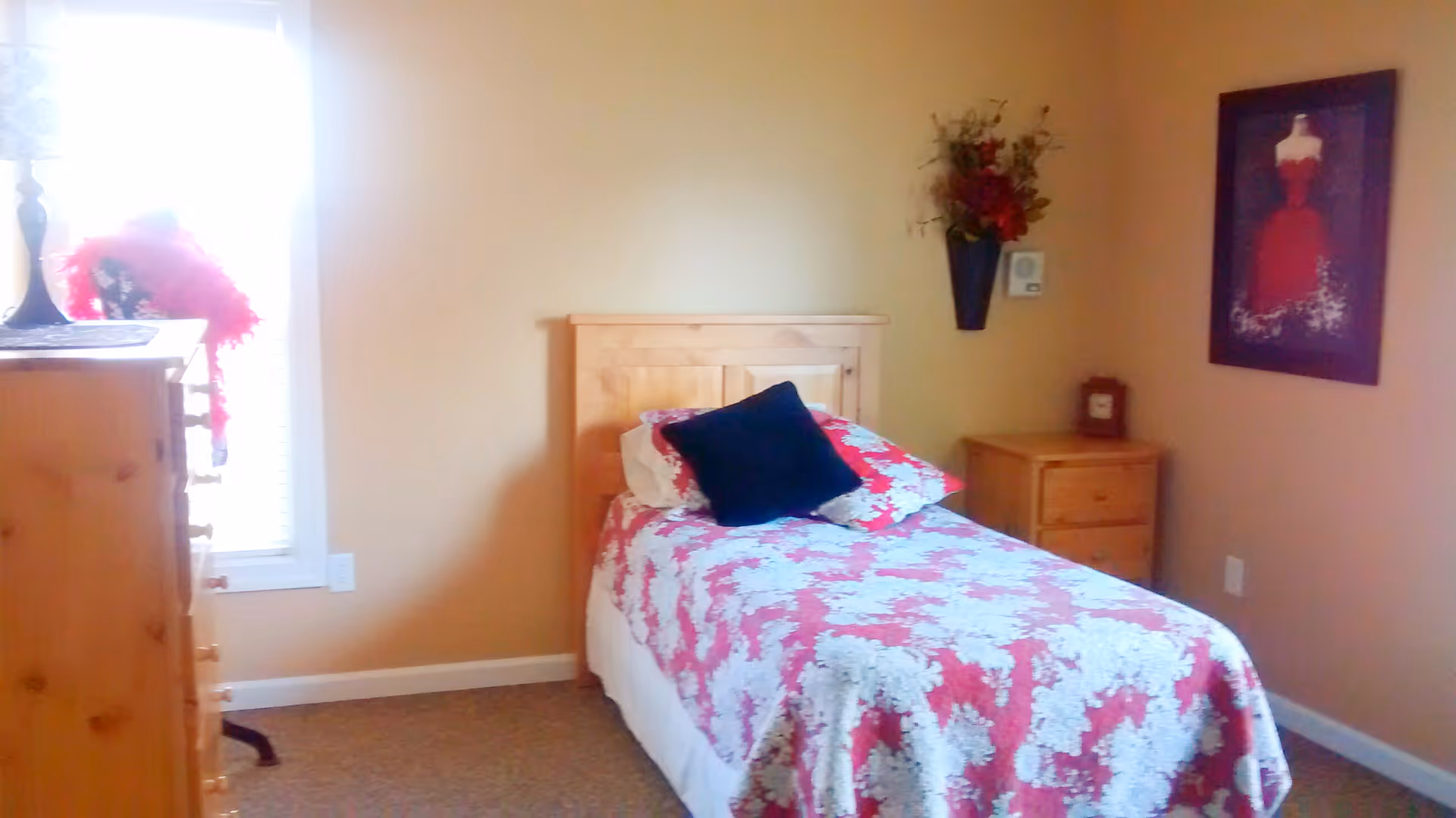 A cozy bedroom with a single wooden bed covered in a red and white floral quilt. There is a black pillow and a floral pillow on the bed. To the right of the bed is a wooden nightstand with a small clock and a wall-mounted flower vase above it. On the left side of the room, there is a wooden dresser near a window with blinds. A framed picture of a red dress hangs on the wall to the right.