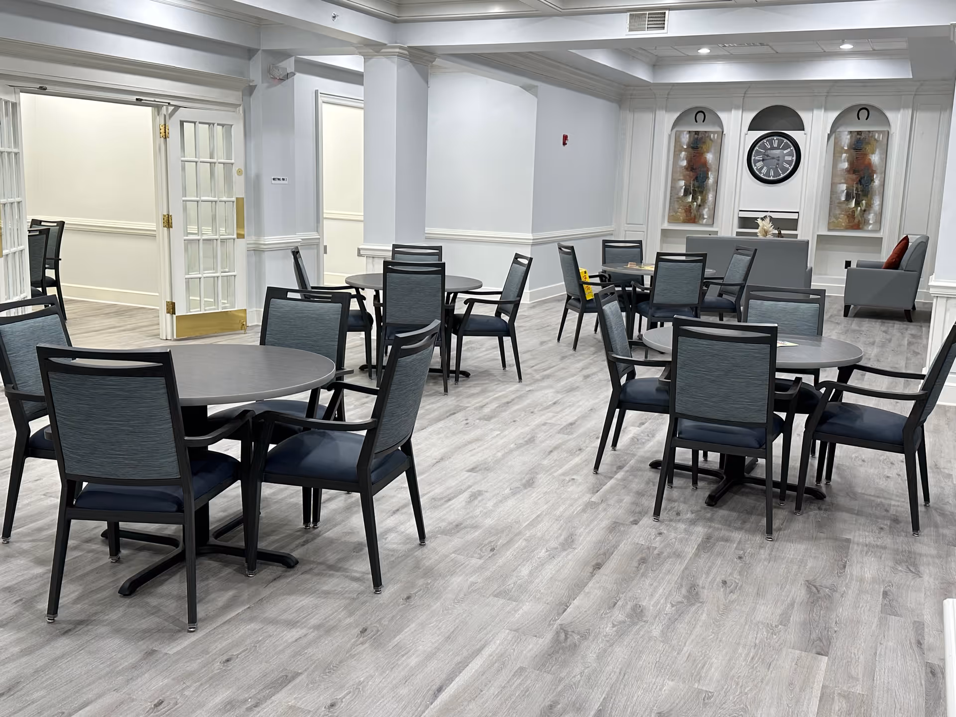 Open dining/activity room with multiple round tables and chairs, light gray wood-look flooring, and a seating area with a clock on the far wall.