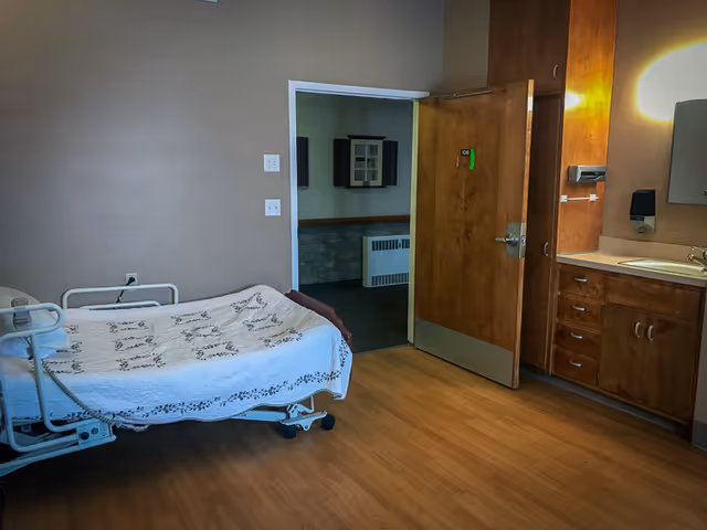 A simple room in a senior living facility with a hospital-style bed covered with a white blanket, wooden flooring, a wooden door slightly open showing another room, and a sink with cabinets and a mirror on the right side.