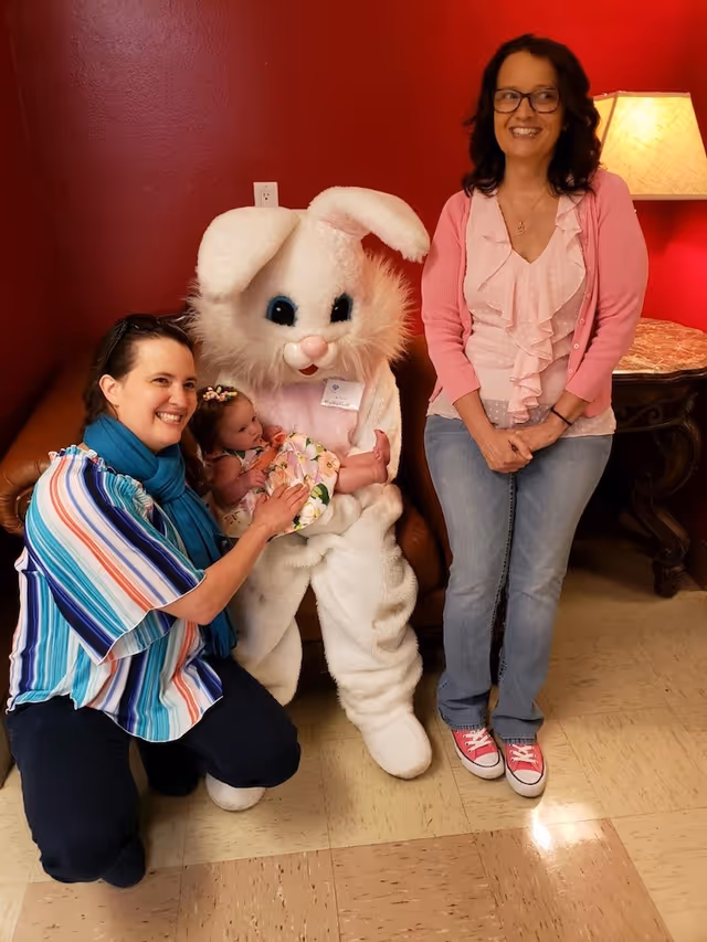 Two women and a baby pose with a person in a bunny costume in a living room area.