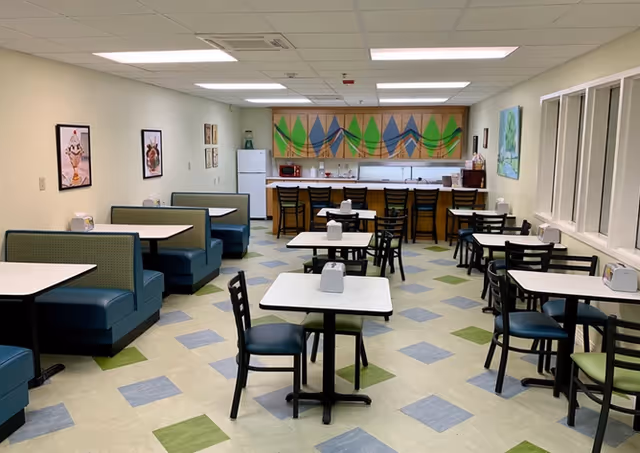 A clean and well-lit dining area with multiple tables and chairs arranged neatly. There are booth-style seats along the left wall and individual tables with chairs in the center and right side of the room. At the back, there is a counter with high chairs, a refrigerator, microwave, and colorful wall art above the counter. The floor has a patterned tile design in blue, green, and beige colors.