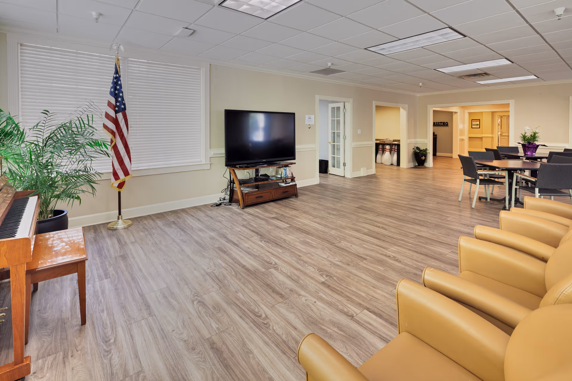 A spacious common area in a senior living facility featuring a row of yellow armchairs, a flat-screen TV on a wooden stand, a piano with a bench, an American flag, a potted plant, and a dining table with chairs. The room has wood flooring, beige walls, and a ceiling with recessed lighting. In the background, there is a hallway with a sign that reads 'Bistro' and some bowling pins visible.