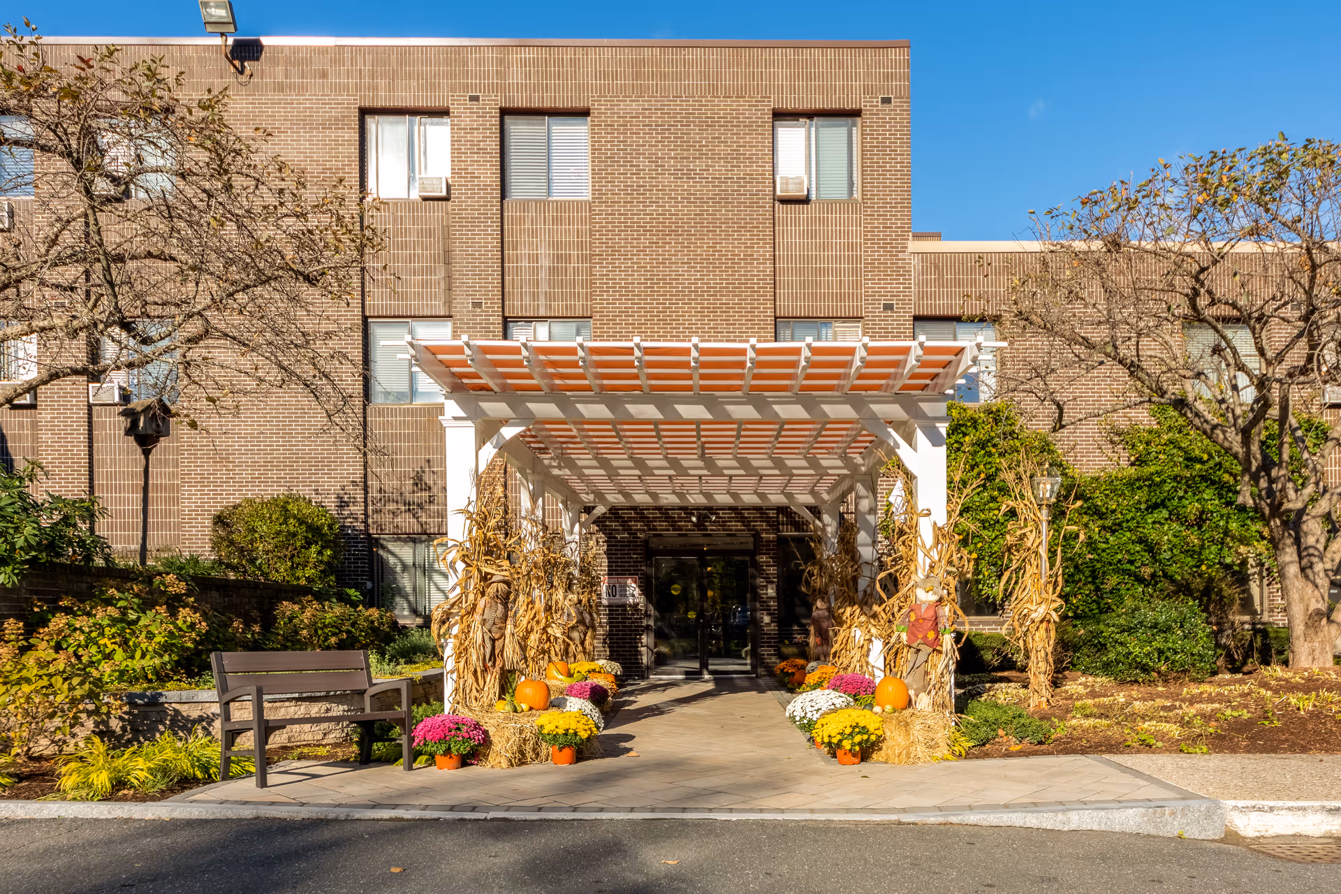 Entrance of a brick building with a white pergola decorated with autumn-themed items including pumpkins, hay bales, corn stalks, and colorful potted flowers. There is a bench to the left and trees and shrubs surrounding the entrance under a clear blue sky.