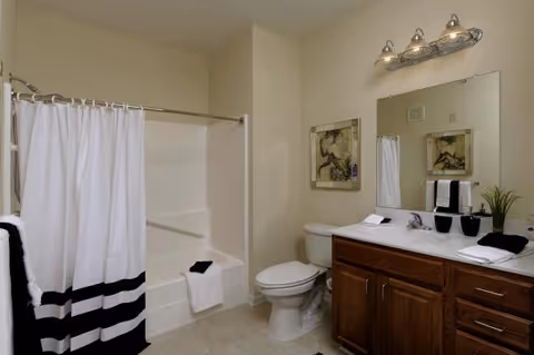 Well-lit bathroom with a tub/shower behind a white-and-black curtain, toilet, and wooden vanity with mirror and light fixture.