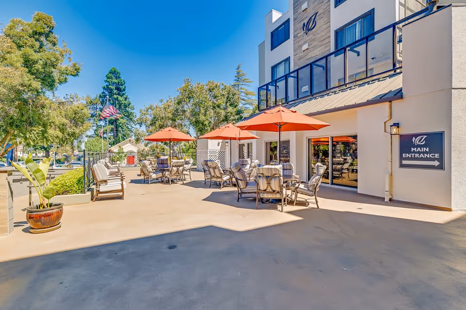 Outdoor patio area at Burlingame Senior Living with several tables and chairs under orange umbrellas, potted plants, trees, and a clear blue sky. A sign on the building indicates the main entrance.