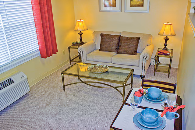 Small living room with a beige loveseat, glass coffee table, side tables with lamps, and a small dining set with blue dishes.