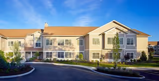 Exterior view of a two-story senior living facility building with beige siding and a brown roof under a clear blue sky. The building is surrounded by landscaped greenery and a paved driveway in front.