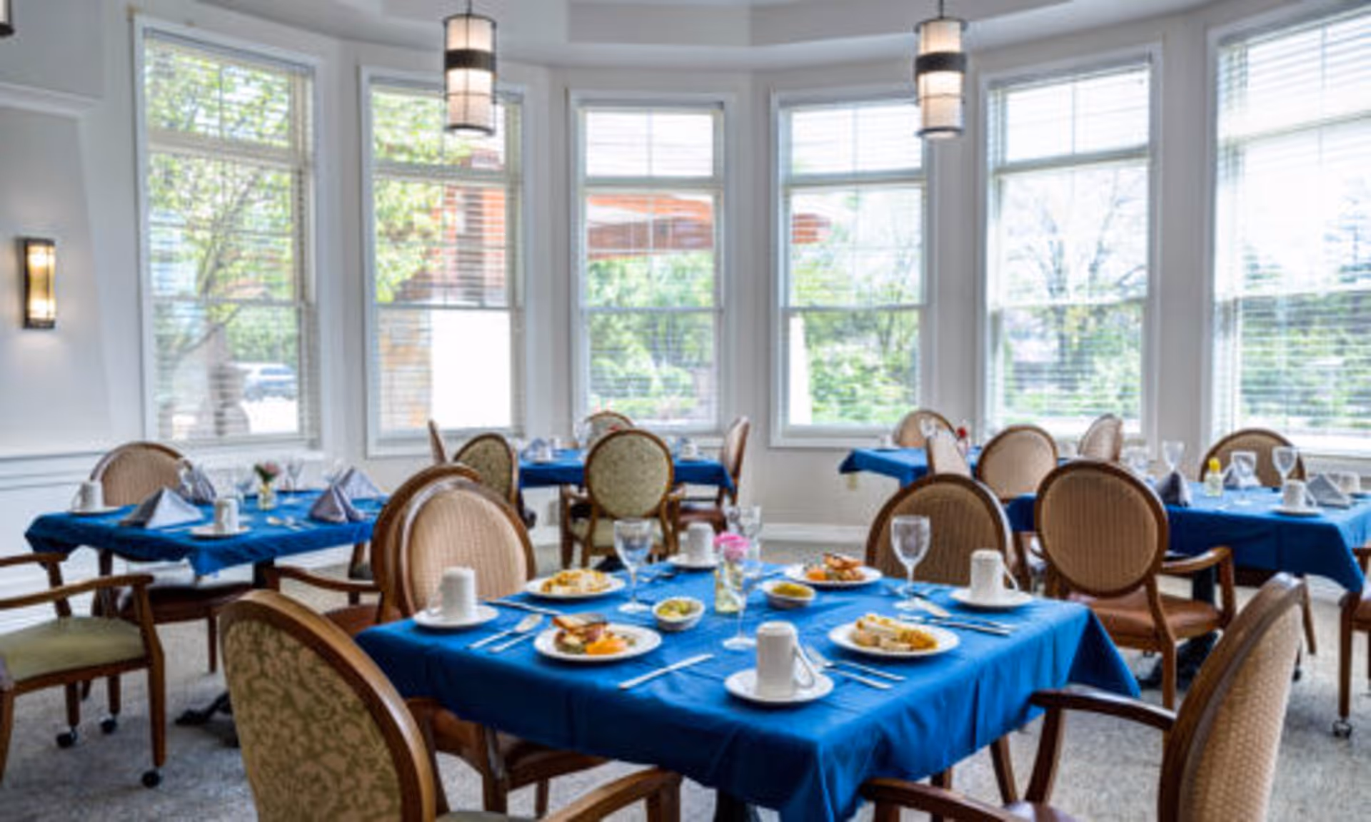 Sunlit dining room with round tables covered in blue tablecloths set with plates, glasses and upholstered chairs in front of large windows.