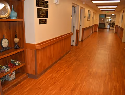 A long, clean hallway with wooden flooring and wood paneling on the lower half of the walls. There is a wooden shelf on the left side displaying decorative items including clocks and vases. Signs on the wall indicate directions to the elevator, admissions office, business office, and nurses station. Doors line the hallway, and the area is well-lit with ceiling lights.
