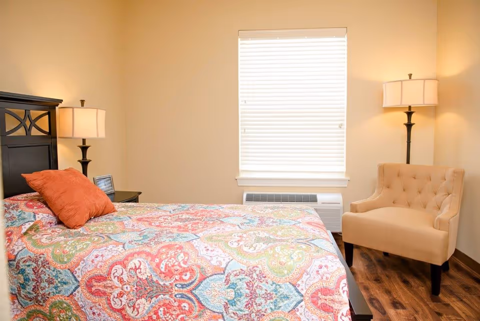 A cozy bedroom with a bed covered in a colorful patterned quilt and an orange pillow. There is a wooden nightstand with a lamp and a small picture frame next to the bed. A beige upholstered armchair is placed near a window with closed blinds. The room has warm beige walls and wooden flooring.