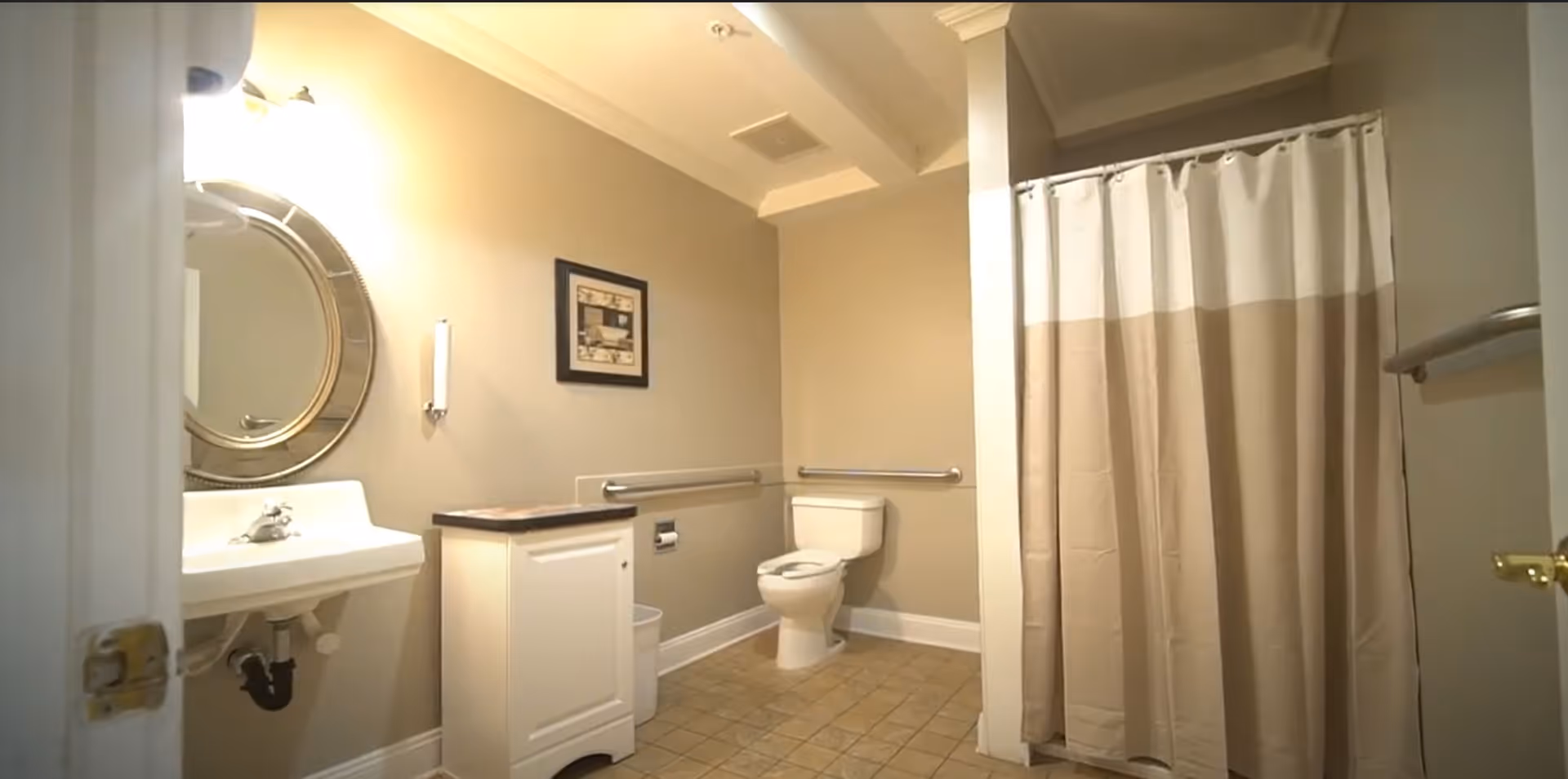 A clean and accessible bathroom with beige walls and tiled floor. The room features a white toilet with grab bars on the walls, a white sink with a round mirror above it, a small cabinet with a dark countertop, and a shower area with a beige and white shower curtain.