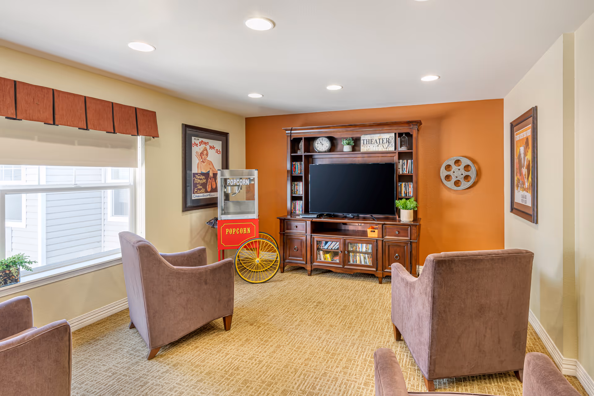 A cozy entertainment room with a large flat-screen TV on a wooden cabinet filled with DVDs. The room has beige carpet, light yellow and orange walls, and three brown upholstered chairs facing the TV. There is a vintage-style popcorn machine with a red and yellow cart, movie posters on the walls, and a decorative film reel mounted on the orange wall.