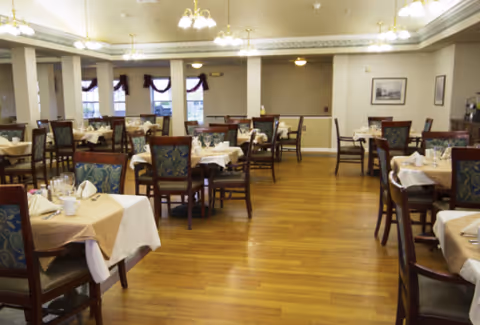 Spacious dining room with multiple neatly set tables, upholstered chairs, hardwood floors, and ceiling light fixtures.