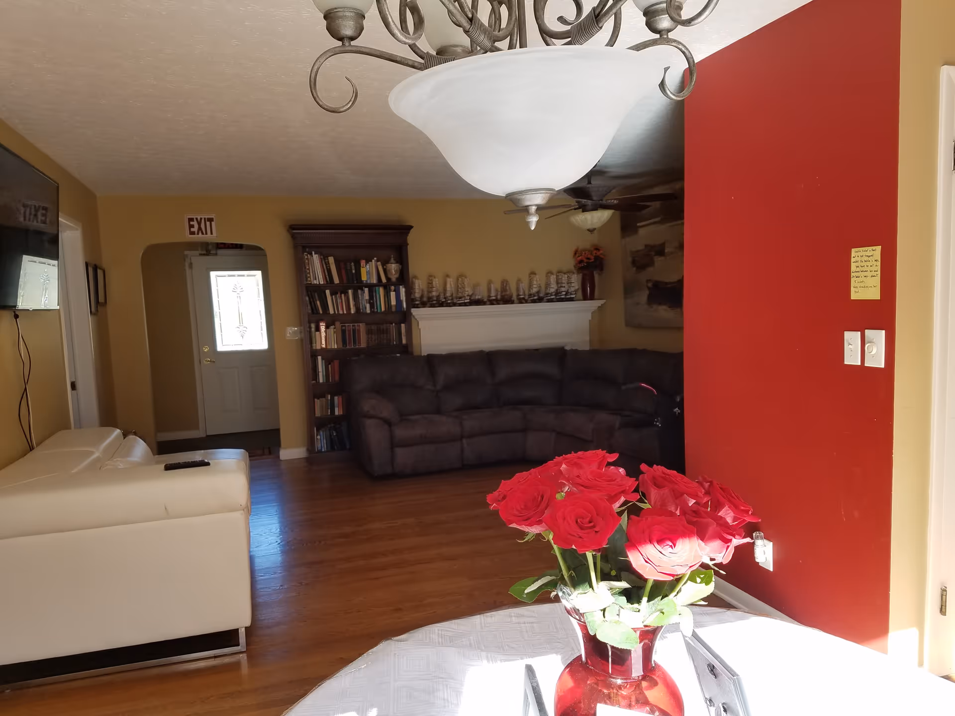 Interior view of a living room with wooden flooring, a white leather sofa on the left, a dark brown sectional sofa against the far wall, a bookshelf filled with books, and a white fireplace mantel decorated with model ships. A red wall is on the right side with a light switch and a small note attached. A glass vase with red roses is placed on a table in the foreground. A ceiling light fixture is visible at the top center.
