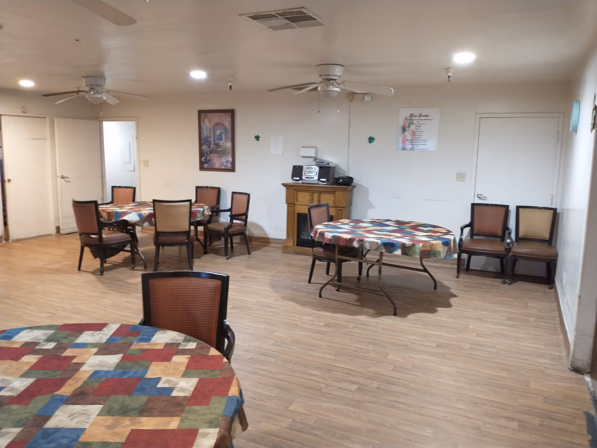A room with several round tables covered with colorful patchwork tablecloths and surrounded by chairs. The room has wooden flooring, white walls, ceiling fans, and recessed lighting. There is a wooden cabinet against the wall with a stereo system on top, and a framed picture and a sign on the wall.