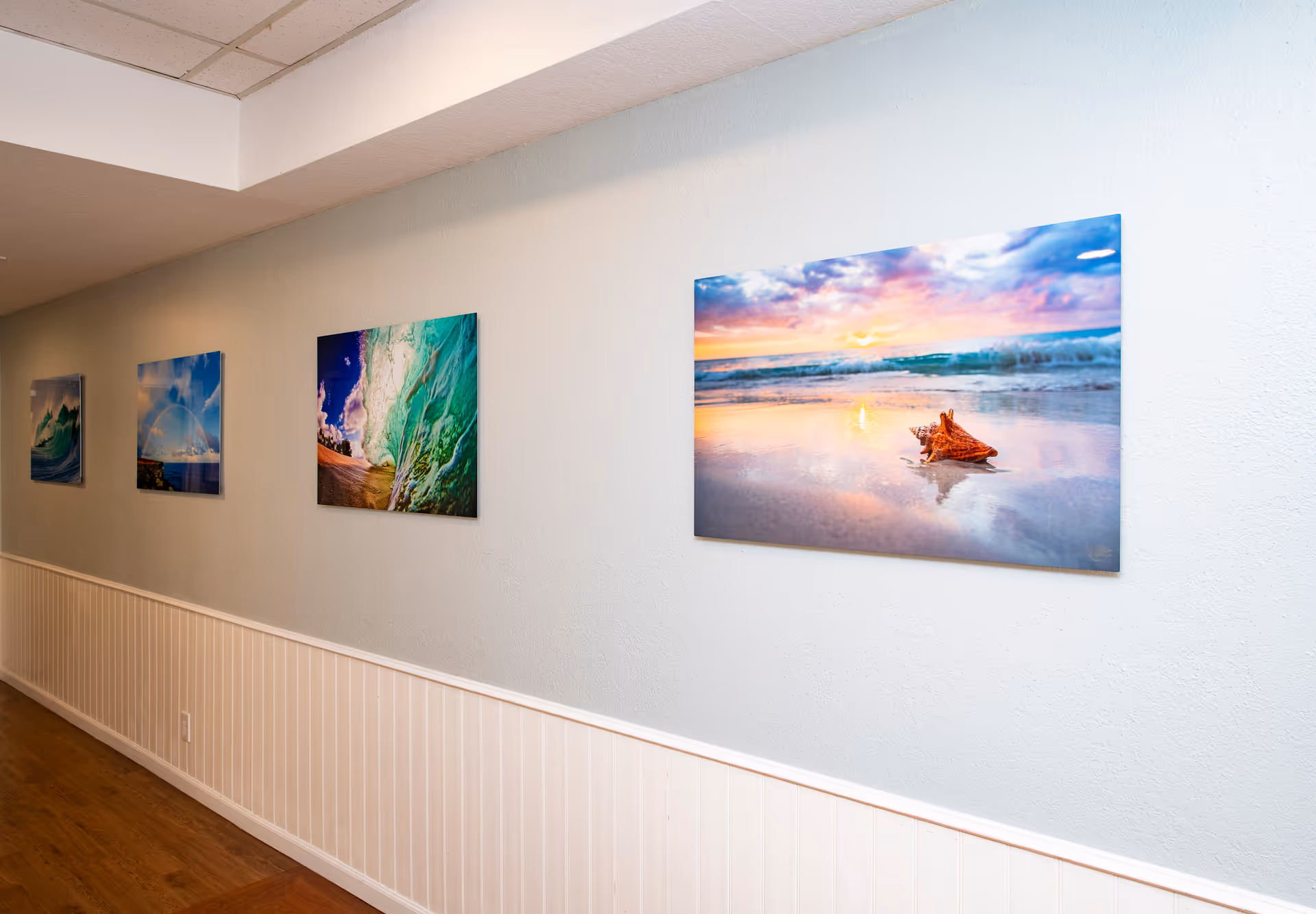 A hallway interior with several framed beach and ocean photographs hung on a light-colored wall above white wainscoting and wood flooring.
