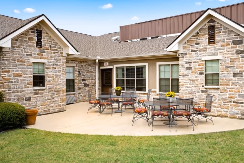 Outdoor patio area at Village on the Park Stonebridge Ranch with stone exterior walls, multiple metal tables and chairs with red cushions, potted yellow flowers on the tables, and a grassy lawn in the foreground under a blue sky with a few clouds.