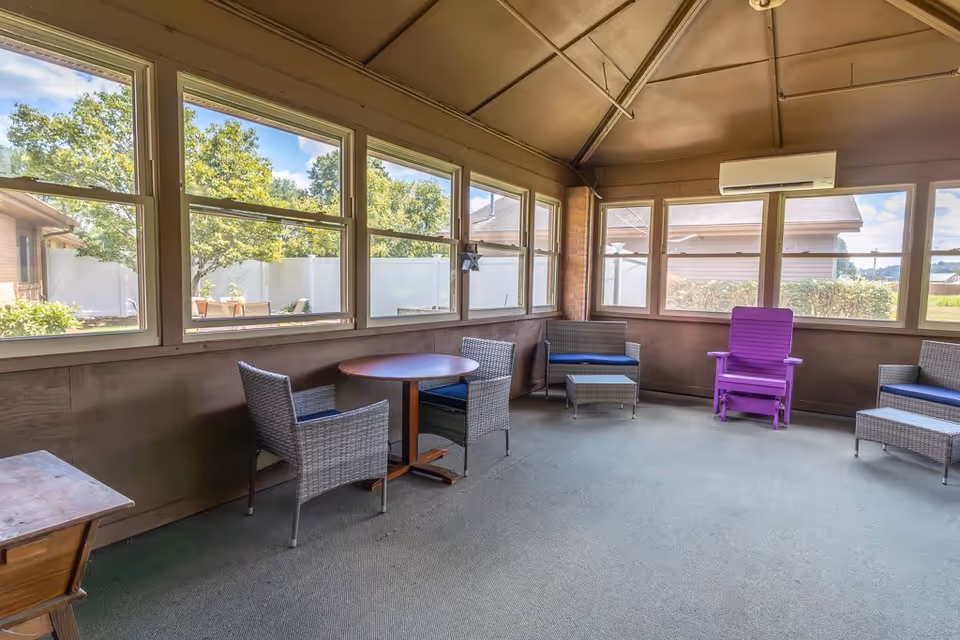 A screened-in porch area with multiple windows showing an outdoor view with trees and a white fence. Inside, there are several pieces of wicker furniture including chairs and a small table, along with a distinctive purple wooden chair. The ceiling is slanted with visible beams and an air conditioning unit mounted on the wall.
