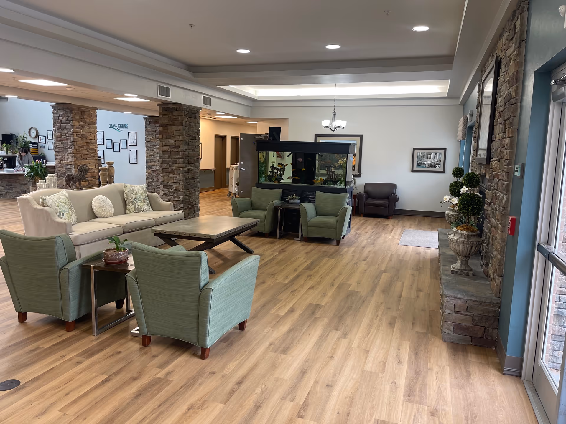 A spacious senior living facility common area with wooden flooring, a beige sofa with decorative pillows, two green armchairs, a wooden coffee table, and a large fish tank. Stone pillars and a reception desk are visible in the background, along with framed pictures on the walls and potted plants near the entrance.