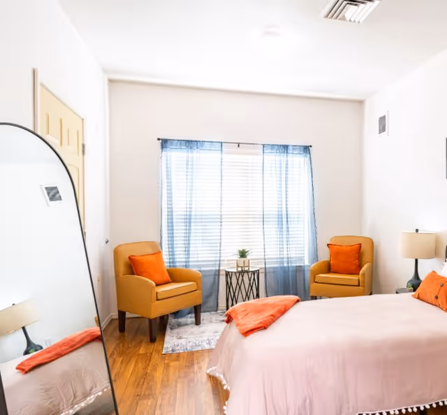 Bright bedroom with a light-pink bed, two mustard armchairs with orange pillows, a full-length mirror, and a window with blue curtains.