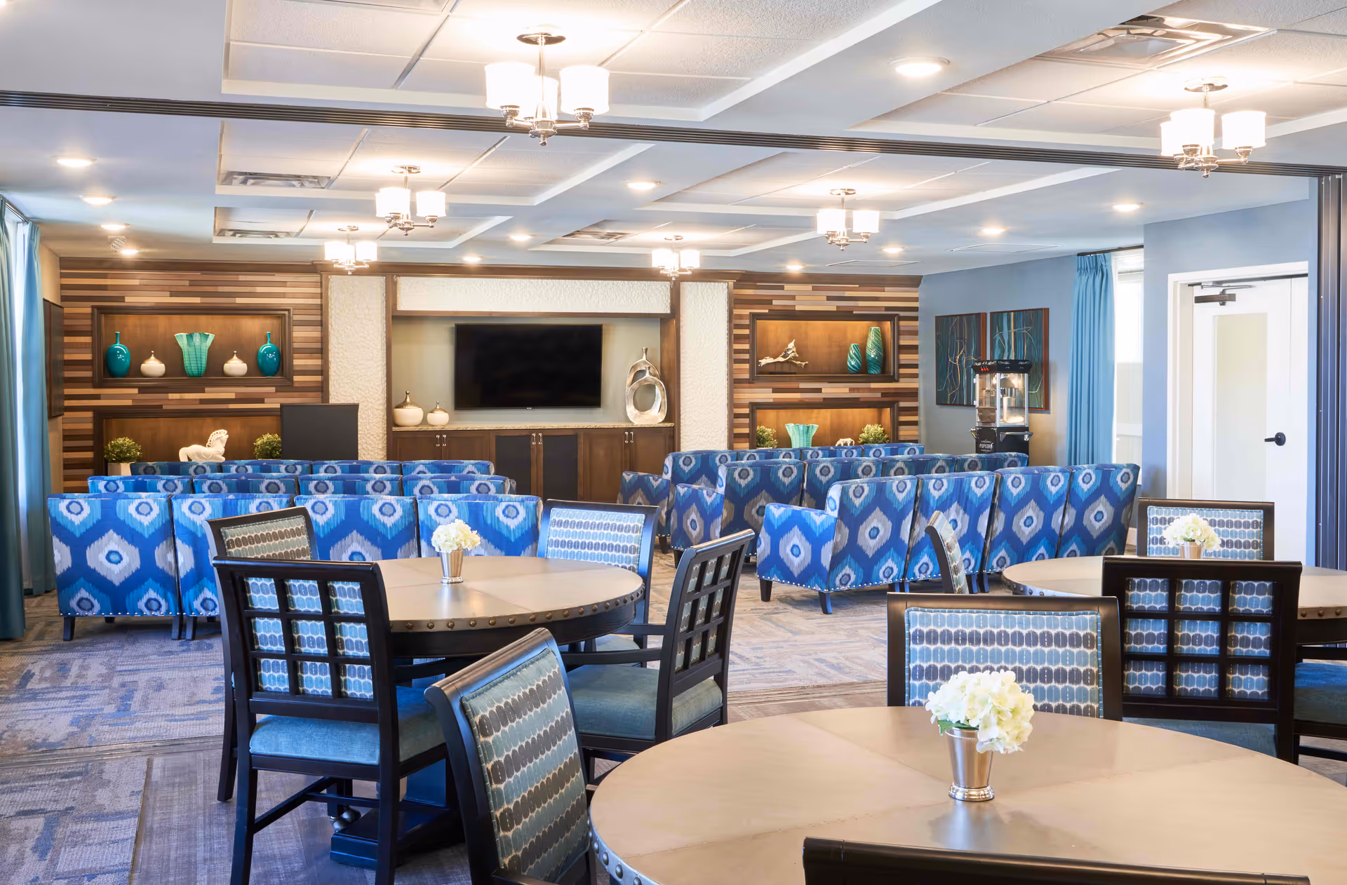 A bright and spacious senior living common area with round tables and chairs in the foreground, decorated with small flower arrangements. In the background, there are blue patterned sofas arranged facing a large flat-screen TV mounted on a wooden accent wall with decorative vases and sculptures. The room has a modern ceiling with recessed lighting and hanging light fixtures.