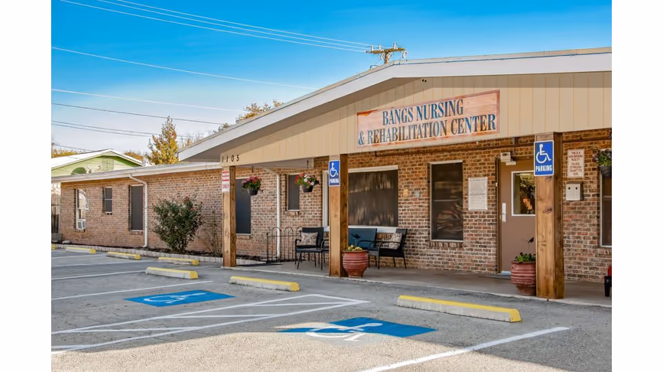 Exterior view of Bangs Nursing and Rehabilitation Center showing a brick building with a covered entrance supported by wooden posts. There are two handicap parking spaces in front, hanging flower pots, and benches near the entrance under a clear blue sky.