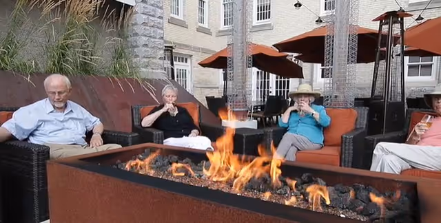 Four elderly people sitting on outdoor wicker furniture with orange cushions around a rectangular fire pit with flames. The setting is an outdoor patio area with tall patio heaters and large orange umbrellas, adjacent to a light-colored brick building with multiple windows.