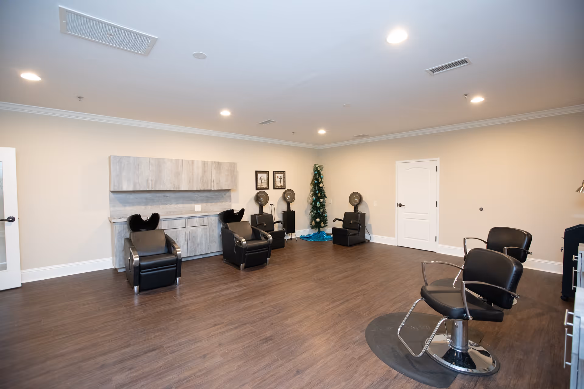 Interior of a salon room in The Timberley facility featuring several black salon chairs, hair drying stations, wooden flooring, beige walls, and a small decorated Christmas tree in the corner.