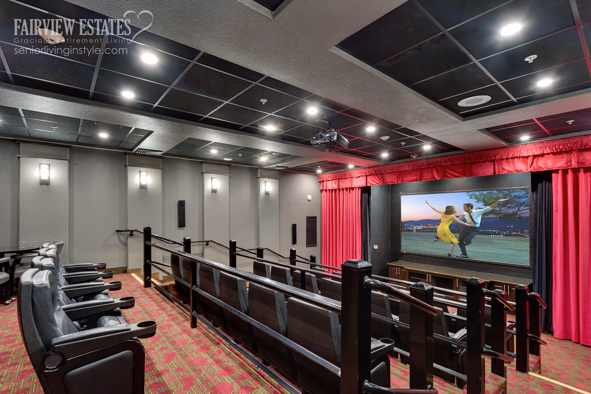A small theater-style screening room with tiered rows of seating, red curtains and a large screen showing a dancing couple.