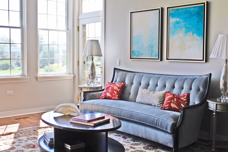 A bright living room with a light blue tufted sofa adorned with two red floral pillows and one gray patterned pillow. Two abstract paintings with blue and beige tones hang on the wall above the sofa. There are two silver table lamps with white shades on round side tables on either side of the sofa. A dark wooden coffee table with books and a decorative item sits on a patterned rug. Large windows and a glass door allow natural light to fill the room.